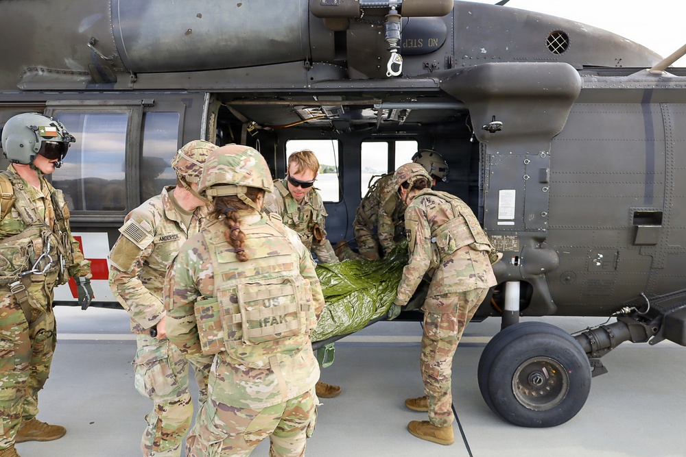Military personnel in U.S. Army OCP (Operational Camouflage Pattern) uniforms are loading or unloading a casualty on a litter into a UH-60 Black Hawk helicopter. The scene appears to be a medical evacuation (MEDEVAC) training exercise or actual casualty transport operation on a tarmac or airfield. A red cross medical bag is visible on the left side, indicating medical personnel involvement.