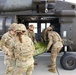 Military personnel in U.S. Army OCP (Operational Camouflage Pattern) uniforms are loading or unloading a casualty on a litter into a UH-60 Black Hawk helicopter. The scene appears to be a medical evacuation (MEDEVAC) training exercise or actual casualty transport operation on a tarmac or airfield. A red cross medical bag is visible on the left side, indicating medical personnel involvement.