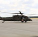 A military medical evacuation (MEDEVAC) helicopter, identified as a UH-60 Black Hawk with tail number 162, is taxiing on a concrete airfield apron with its rotors spinning. The aircraft is painted in olive drab and bears a prominent red cross emblem on its fuselage, indicating its medical role. A second military aircraft is partially visible in the background to the left.