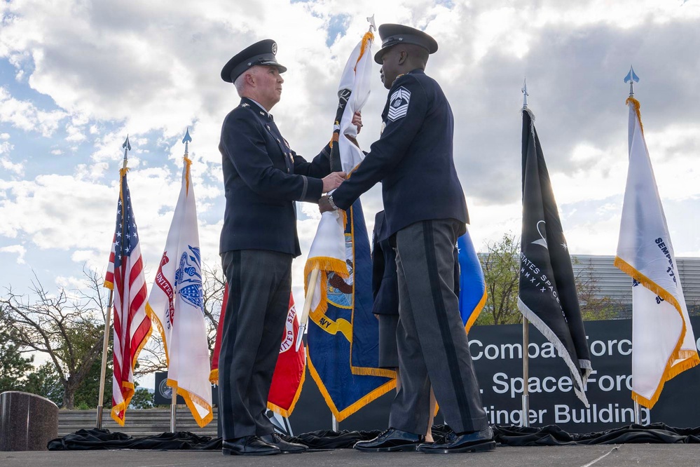 Two military personnel in dress uniforms are participating in a formal flag ceremony, likely a change of command or activation ceremony. They are handling a white flag together on an outdoor stage surrounded by multiple military service flags. A sign in the background references 'Combat Force,' 'Space Force,' and a building name ending in 'tinger Building.'