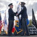 Two military personnel in dress uniforms are participating in a formal flag ceremony, likely a change of command or activation ceremony. They are handling a white flag together on an outdoor stage surrounded by multiple military service flags. A sign in the background references 'Combat Force,' 'Space Force,' and a building name ending in 'tinger Building.'