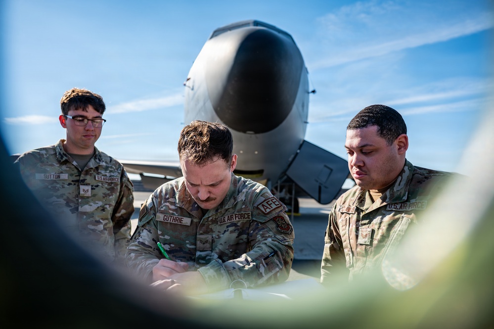 Three U.S. Air Force personnel in OCP (Operational Camouflage Pattern) uniforms are gathered on a flight line, with the center individual writing on a document or form. They are positioned in front of a large military aircraft, identifiable by its distinctive nose cone visible in the background. The scene appears to depict a pre-flight inspection, maintenance documentation, or operational checklist review.
