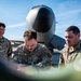 Three U.S. Air Force personnel in OCP (Operational Camouflage Pattern) uniforms are gathered on a flight line, with the center individual writing on a document or form. They are positioned in front of a large military aircraft, identifiable by its distinctive nose cone visible in the background. The scene appears to depict a pre-flight inspection, maintenance documentation, or operational checklist review.