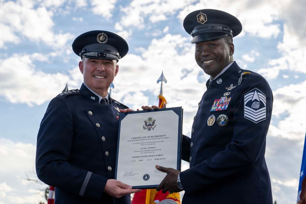 Two United States Space Force personnel in dress uniforms are posing together while holding a Certificate of Retirement. The certificate is issued to Jacob C. Simmons, retired from the United States Space Force on the first day of February 2027. The ceremony appears to be taking place outdoors with military flags visible in the background.