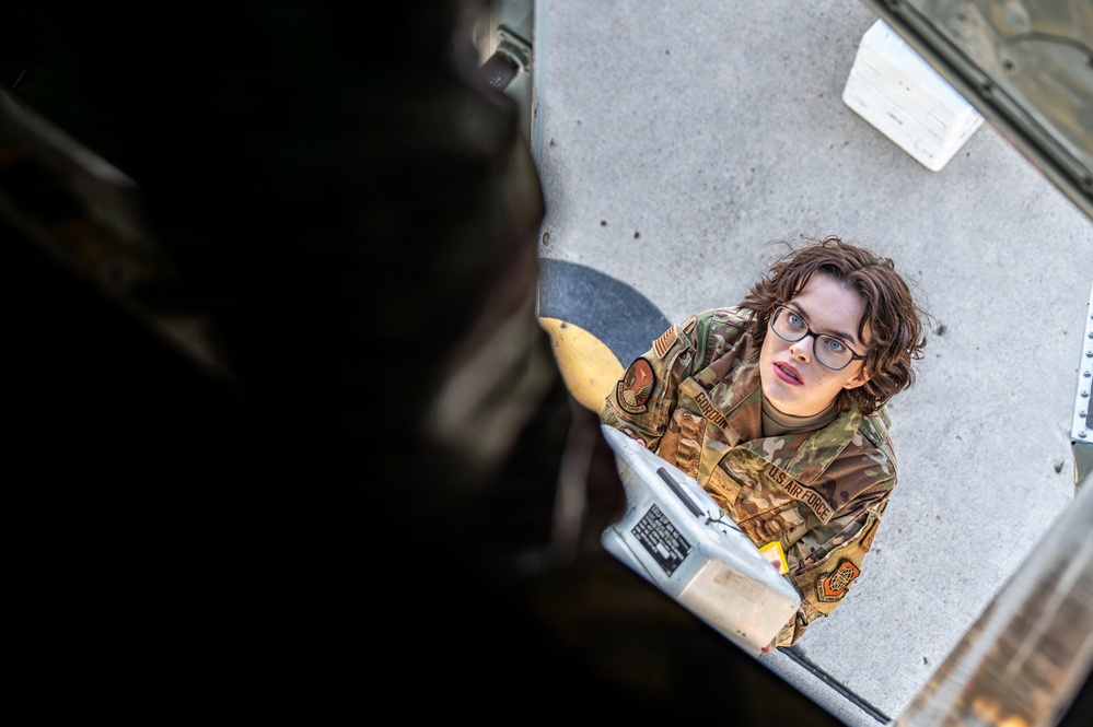 A U.S. Air Force service member in OCP (Operational Camouflage Pattern) uniform is photographed from an elevated angle, appearing to look upward toward the camera. The individual is wearing glasses and has a name tape and 'U.S. AIR FORCE' tape visible on their uniform. They appear to be working on or inspecting military equipment, possibly an aircraft or vehicle component.