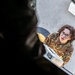 A U.S. Air Force service member in OCP (Operational Camouflage Pattern) uniform is photographed from an elevated angle, appearing to look upward toward the camera. The individual is wearing glasses and has a name tape and 'U.S. AIR FORCE' tape visible on their uniform. They appear to be working on or inspecting military equipment, possibly an aircraft or vehicle component.