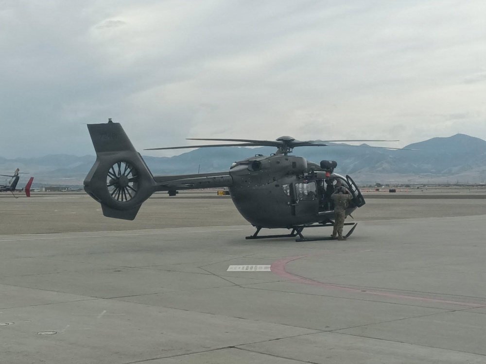 A military helicopter, consistent with the UH-72 Lakota (Airbus H145) design, is parked on a tarmac at what appears to be a military airfield. At least one soldier in camouflage uniform is visible near the aircraft's cabin door. Mountains and an overcast sky are visible in the background.