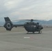A military helicopter, consistent with the UH-72 Lakota (Airbus H145) design, is parked on a tarmac at what appears to be a military airfield. At least one soldier in camouflage uniform is visible near the aircraft's cabin door. Mountains and an overcast sky are visible in the background.