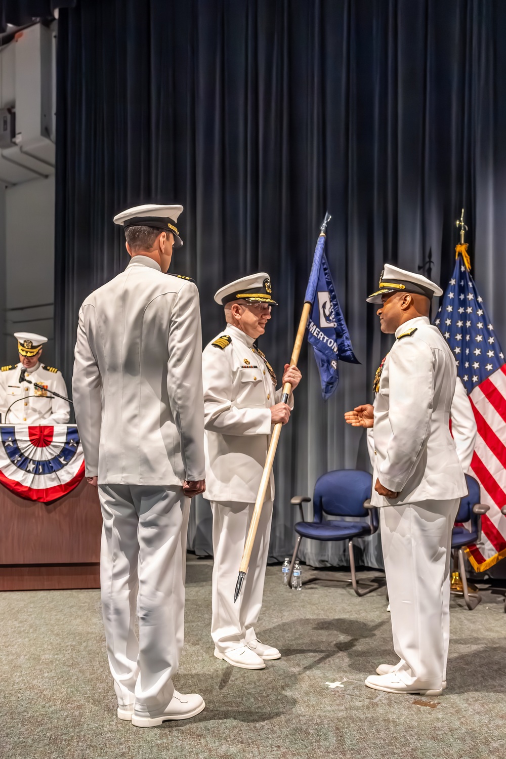 A formal U.S. Navy change of command ceremony is taking place on a stage. A central officer is holding a unit guidon (flag) in what appears to be the traditional passing of the colors between an outgoing and incoming commanding officer. The setting includes patriotic decorations, an American flag, and a presiding officer at a podium in the background.