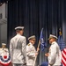 A formal U.S. Navy change of command ceremony is taking place on a stage. A central officer is holding a unit guidon (flag) in what appears to be the traditional passing of the colors between an outgoing and incoming commanding officer. The setting includes patriotic decorations, an American flag, and a presiding officer at a podium in the background.
