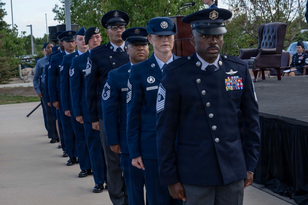 A formation of U.S. Air Force and Space Force personnel in dress blue uniforms are standing at attention during a formal military ceremony. The group is arranged in a single-file diagonal line in front of a stage, with varying ranks visible including senior NCOs and at least one Chief Master Sergeant. A podium and ceremonial chair are visible on the stage in the background.