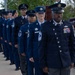 A formation of U.S. Air Force and Space Force personnel in dress blue uniforms are standing at attention during a formal military ceremony. The group is arranged in a single-file diagonal line in front of a stage, with varying ranks visible including senior NCOs and at least one Chief Master Sergeant. A podium and ceremonial chair are visible on the stage in the background.