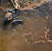 A scuba diver in full black wetsuit and diving gear is partially surfaced in murky, debris-filled brown water, photographed from above. The water contains numerous pieces of dead reeds, sticks, and organic debris floating on the surface. The diver appears to be conducting an underwater search or inspection operation near a structure.
