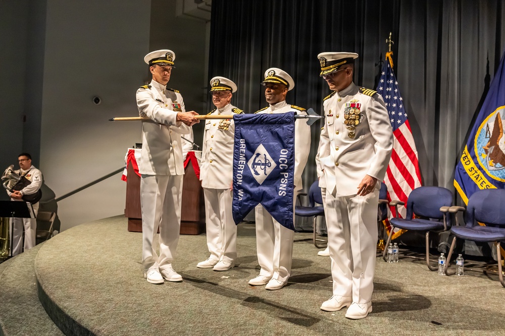 A formal U.S. Navy change of command ceremony is taking place on a stage, with officers in dress white uniforms participating in the transfer of a unit guidon. The pennant visible reads 'OICC PSNS Bremerton, WA,' indicating this is the Officer in Charge of Construction, Puget Sound Naval Shipyard, located in Bremerton, Washington. Four naval officers are centrally involved in the ceremony, with an American flag and a U.S. Navy flag visible in the background.