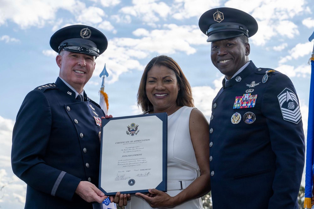 Two military officers in dress uniforms are presenting a Certificate of Appreciation to a civilian woman at what appears to be an official military ceremony. The certificate is made out to 'Analida Simmons' and is dated February 2022. All three individuals are smiling and posing for the photograph outdoors.