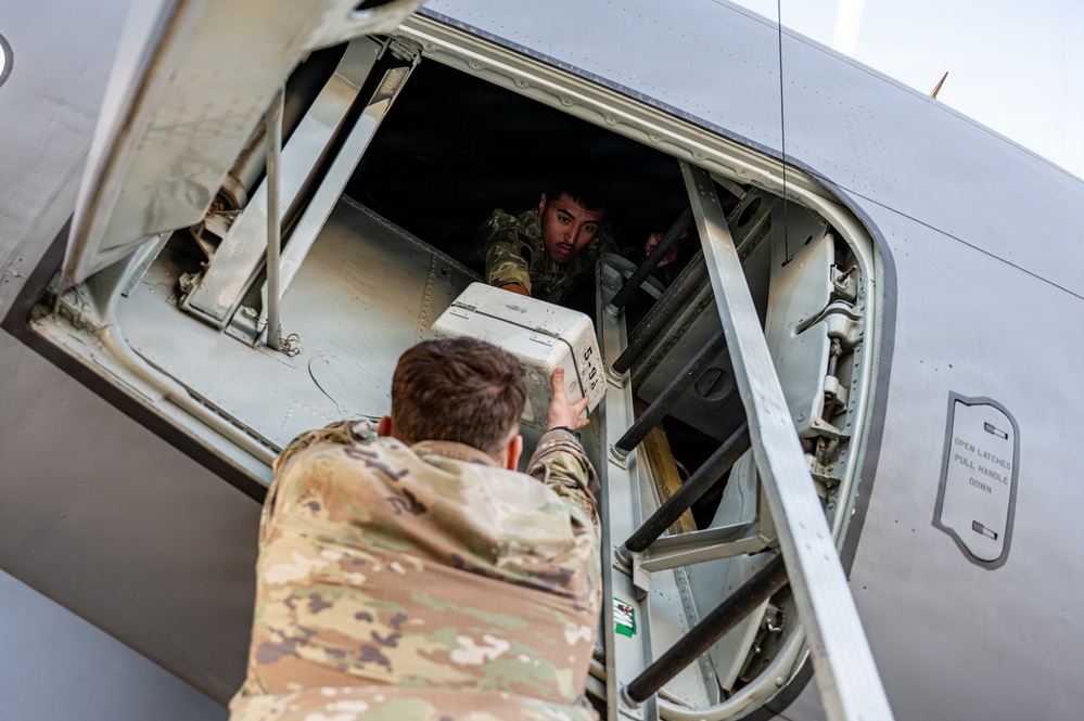 Two military personnel in MultiCam/OCP camouflage uniforms are loading cargo into a military aircraft through a side door or hatch. One service member is outside the aircraft passing a white container upward, while a second is inside the aircraft receiving it. The aircraft appears to be a large military transport, identifiable by its gray fuselage and structural framing.