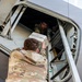 Two military personnel in MultiCam/OCP camouflage uniforms are loading cargo into a military aircraft through a side door or hatch. One service member is outside the aircraft passing a white container upward, while a second is inside the aircraft receiving it. The aircraft appears to be a large military transport, identifiable by its gray fuselage and structural framing.