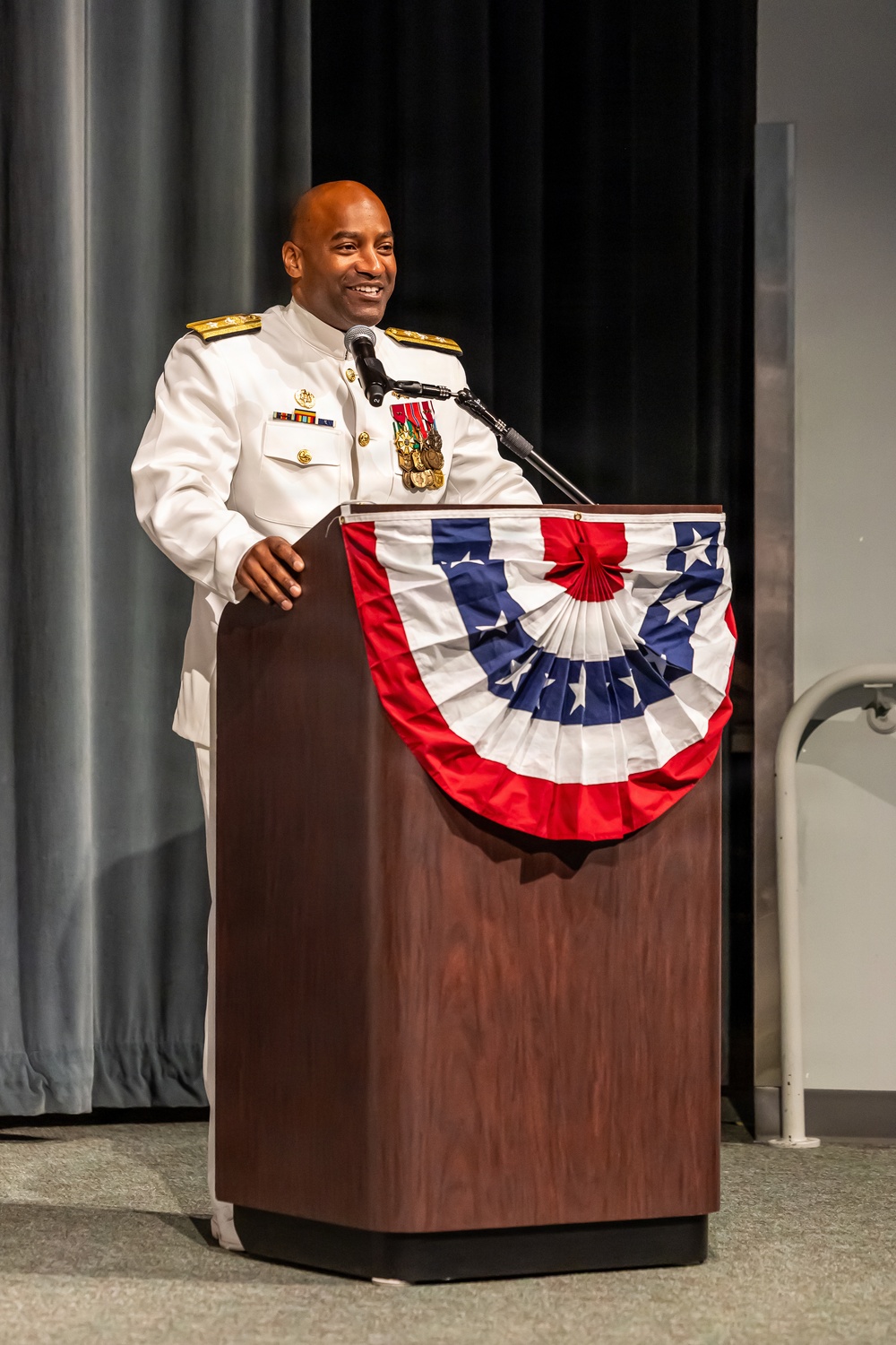 A senior military officer in white dress uniform stands at a wooden podium decorated with a patriotic red, white, and blue bunting. He is smiling and speaking into a microphone at what appears to be a formal military ceremony or official event. His uniform displays multiple medals, ribbons, and gold shoulder boards indicating high rank.