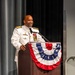 A senior military officer in white dress uniform stands at a wooden podium decorated with a patriotic red, white, and blue bunting. He is smiling and speaking into a microphone at what appears to be a formal military ceremony or official event. His uniform displays multiple medals, ribbons, and gold shoulder boards indicating high rank.