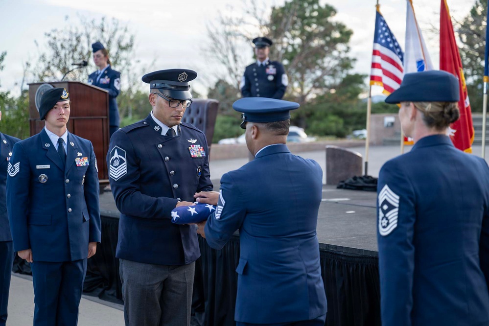 A formal U.S. Air Force military ceremony is taking place outdoors, likely a retirement or memorial service. A senior enlisted member (Chief Master Sergeant) is receiving a folded American flag from another uniformed officer, a traditional military honor. Multiple service members in dress blues are present, with flags and a podium visible in the background.