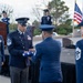 A formal U.S. Air Force military ceremony is taking place outdoors, likely a retirement or memorial service. A senior enlisted member (Chief Master Sergeant) is receiving a folded American flag from another uniformed officer, a traditional military honor. Multiple service members in dress blues are present, with flags and a podium visible in the background.