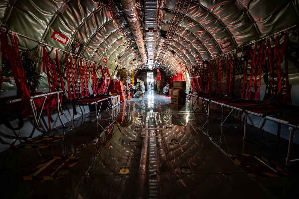 The image shows the interior of a large military transport aircraft, likely a C-17 or similar airlifter, configured for aeromedical evacuation or cargo transport. The fuselage interior features distinctive red litter/stretcher stanchions mounted along both sides of the curved walls, with reflective insulation lining. Several personnel are visible mid-cabin, appearing to load or arrange equipment and cargo boxes.