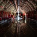 The image shows the interior of a large military transport aircraft, likely a C-17 or similar airlifter, configured for aeromedical evacuation or cargo transport. The fuselage interior features distinctive red litter/stretcher stanchions mounted along both sides of the curved walls, with reflective insulation lining. Several personnel are visible mid-cabin, appearing to load or arrange equipment and cargo boxes.
