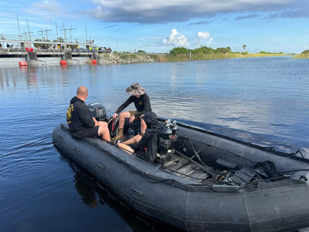 Two individuals are seated in a black inflatable tactical/military-style rigid inflatable boat (RIB) on a calm waterway. The boat is equipped with diving equipment including scuba tanks and an outboard motor. In the background, a water control structure or lock facility is visible with several people standing on it.