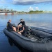 Two individuals are seated in a black inflatable tactical/military-style rigid inflatable boat (RIB) on a calm waterway. The boat is equipped with diving equipment including scuba tanks and an outboard motor. In the background, a water control structure or lock facility is visible with several people standing on it.