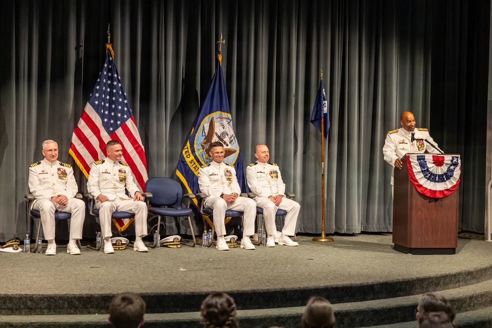 A formal U.S. Navy military ceremony is taking place on a stage, likely a change of command or award ceremony. Five naval officers in white dress uniforms are present, with one speaking at a podium decorated with a patriotic bunting. Three flags are displayed in the background, including the U.S. flag and what appears to be a U.S. Navy command flag.