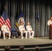 A formal U.S. Navy military ceremony is taking place on a stage, likely a change of command or award ceremony. Five naval officers in white dress uniforms are present, with one speaking at a podium decorated with a patriotic bunting. Three flags are displayed in the background, including the U.S. flag and what appears to be a U.S. Navy command flag.