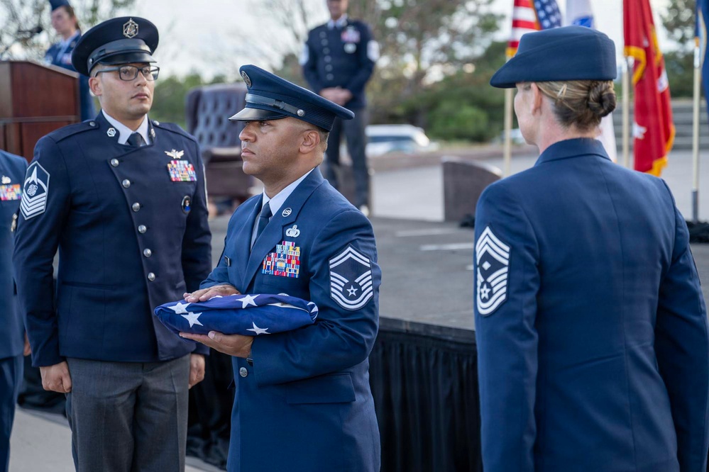 A formal U.S. Air Force military ceremony is taking place outdoors, likely a retirement or change of command ceremony. A senior non-commissioned officer in the center is holding a folded American flag, preparing to present it to a female Chief Master Sergeant facing him. Multiple uniformed personnel with service dress blues and various rank insignia are present on and near a stage.