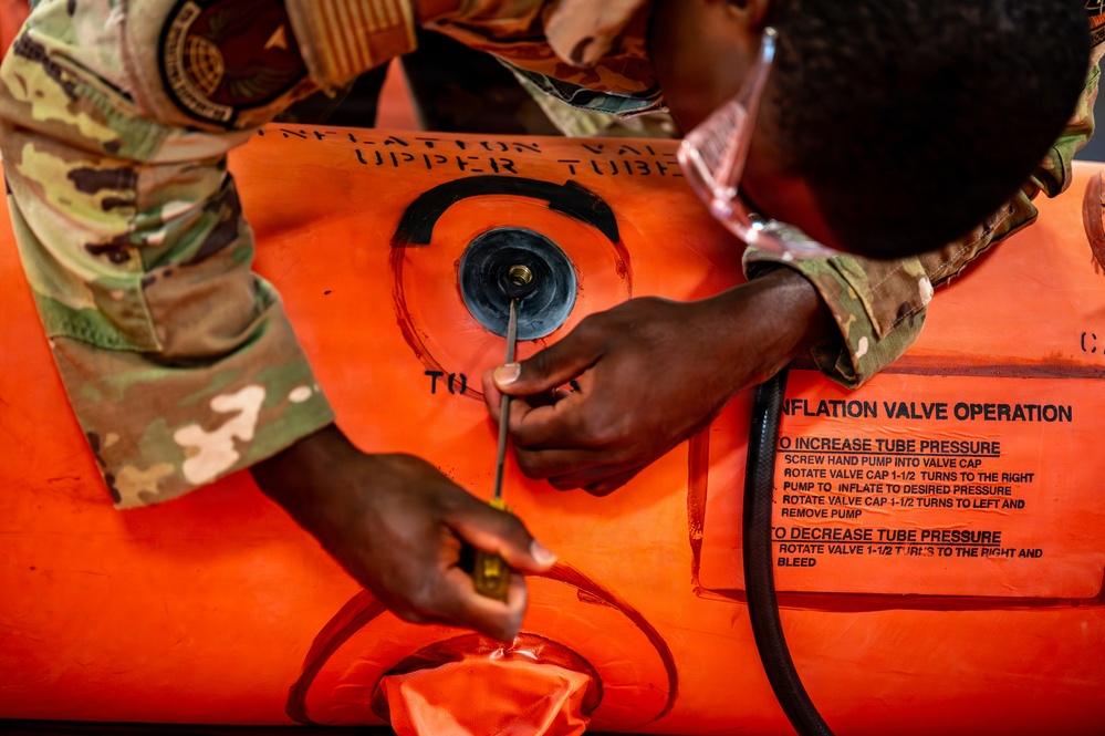 Two individuals in military multicam/OCP camouflage uniforms are working on an orange inflatable raft or survival craft, specifically operating the inflation valve on the upper tube. One person is using a tool, likely a screwdriver or valve key, to adjust the inflation valve. The orange equipment displays clear instructional labels for inflation valve operation.