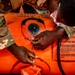 Two individuals in military multicam/OCP camouflage uniforms are working on an orange inflatable raft or survival craft, specifically operating the inflation valve on the upper tube. One person is using a tool, likely a screwdriver or valve key, to adjust the inflation valve. The orange equipment displays clear instructional labels for inflation valve operation.