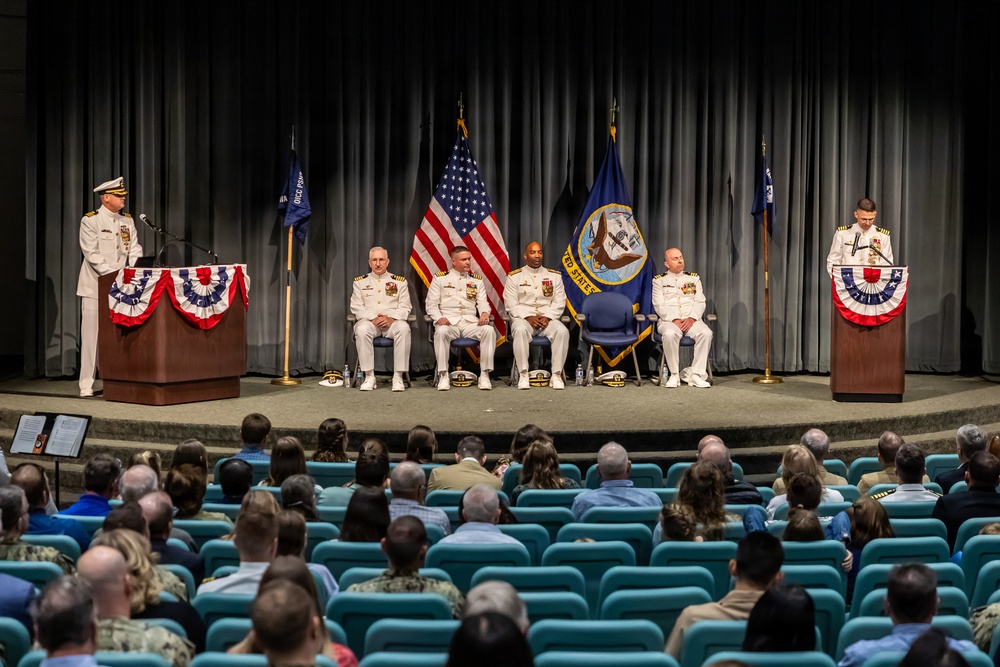 A formal U.S. Navy change of command or retirement ceremony taking place in an auditorium or theater. Four naval officers in white dress uniforms are seated on stage between two podiums, with an officer speaking at the left podium and another standing at the right podium. A sizeable audience is seated in teal auditorium chairs facing the stage.