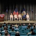 A formal U.S. Navy change of command or retirement ceremony taking place in an auditorium or theater. Four naval officers in white dress uniforms are seated on stage between two podiums, with an officer speaking at the left podium and another standing at the right podium. A sizeable audience is seated in teal auditorium chairs facing the stage.