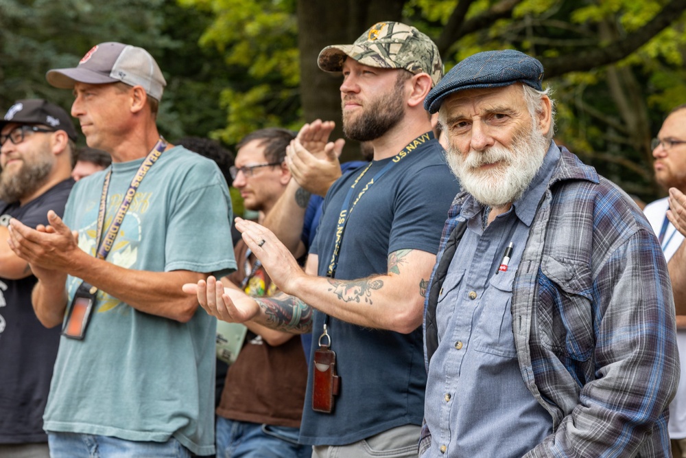 A group of men are gathered outdoors, with several visibly applauding. One man in the foreground wears a U.S. Navy t-shirt and camouflage cap, suggesting a military or veterans-related event. An older man with a white beard and flat cap stands prominently in the right foreground, looking toward the camera.