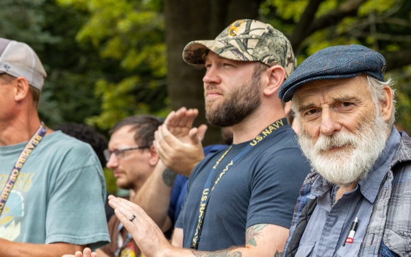 A group of men are gathered outdoors, with several visibly applauding. One man in the foreground wears a U.S. Navy t-shirt and camouflage cap, suggesting a military or veterans-related event. An older man with a white beard and flat cap stands prominently in the right foreground, looking toward the camera.