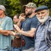 A group of men are gathered outdoors, with several visibly applauding. One man in the foreground wears a U.S. Navy t-shirt and camouflage cap, suggesting a military or veterans-related event. An older man with a white beard and flat cap stands prominently in the right foreground, looking toward the camera.