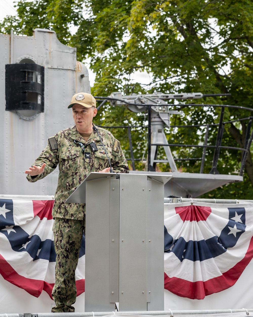 A U.S. Navy officer in NWU Type I (digital camouflage) uniform is delivering a speech at a podium during what appears to be an official military ceremony. The setting appears to be aboard or adjacent to a naval vessel, decorated with patriotic American bunting. The officer's name tape reads 'NICE' and rank insignia indicates a Captain (O-6), with 'U.S. NAVY' branch tape visible.