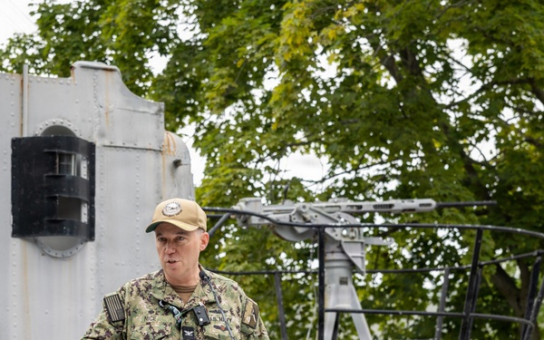 A U.S. Navy officer in NWU Type I (digital camouflage) uniform is delivering a speech at a podium during what appears to be an official military ceremony. The setting appears to be aboard or adjacent to a naval vessel, decorated with patriotic American bunting. The officer's name tape reads 'NICE' and rank insignia indicates a Captain (O-6), with 'U.S. NAVY' branch tape visible.