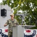 A U.S. Navy officer in NWU Type I (digital camouflage) uniform is delivering a speech at a podium during what appears to be an official military ceremony. The setting appears to be aboard or adjacent to a naval vessel, decorated with patriotic American bunting. The officer's name tape reads 'NICE' and rank insignia indicates a Captain (O-6), with 'U.S. NAVY' branch tape visible.