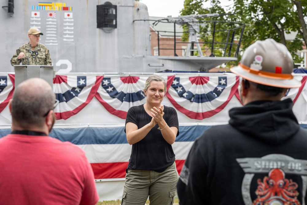 An outdoor military or patriotic ceremony is taking place near what appears to be a naval vessel, decorated with American bunting flags. A woman in a black shirt and cargo pants stands in the foreground clapping, while a uniformed Navy personnel speaks at a podium in the background. Several attendees are visible watching the proceedings.