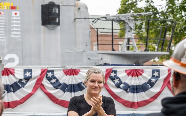 An outdoor military or patriotic ceremony is taking place near what appears to be a naval vessel, decorated with American bunting flags. A woman in a black shirt and cargo pants stands in the foreground clapping, while a uniformed Navy personnel speaks at a podium in the background. Several attendees are visible watching the proceedings.