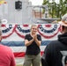 An outdoor military or patriotic ceremony is taking place near what appears to be a naval vessel, decorated with American bunting flags. A woman in a black shirt and cargo pants stands in the foreground clapping, while a uniformed Navy personnel speaks at a podium in the background. Several attendees are visible watching the proceedings.