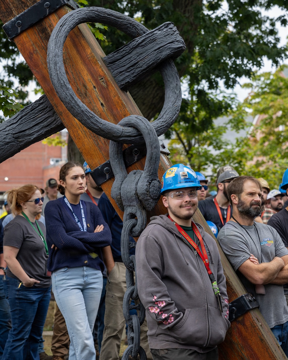 A group of people, many wearing hard hats and lanyards, are gathered outdoors near a large wooden and iron anchor or ship rigging structure. The crowd appears to be attending a maritime heritage or shipyard event, possibly a demonstration or tour. The setting includes trees and brick buildings in the background, suggesting an outdoor museum or naval yard environment.