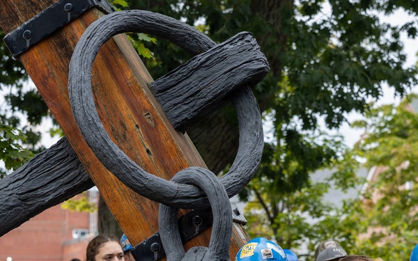 A group of people, many wearing hard hats and lanyards, are gathered outdoors near a large wooden and iron anchor or ship rigging structure. The crowd appears to be attending a maritime heritage or shipyard event, possibly a demonstration or tour. The setting includes trees and brick buildings in the background, suggesting an outdoor museum or naval yard environment.