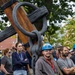 A group of people, many wearing hard hats and lanyards, are gathered outdoors near a large wooden and iron anchor or ship rigging structure. The crowd appears to be attending a maritime heritage or shipyard event, possibly a demonstration or tour. The setting includes trees and brick buildings in the background, suggesting an outdoor museum or naval yard environment.