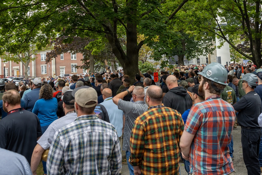 A large outdoor crowd of predominantly adult males has gathered in what appears to be a public square or plaza near brick institutional buildings. One individual in the foreground right is wearing a white hard hat marked with the number '712', suggesting union or trade worker affiliation. The crowd appears to be attending a rally, demonstration, or public gathering, with people facing toward a central focal point obscured by trees.
