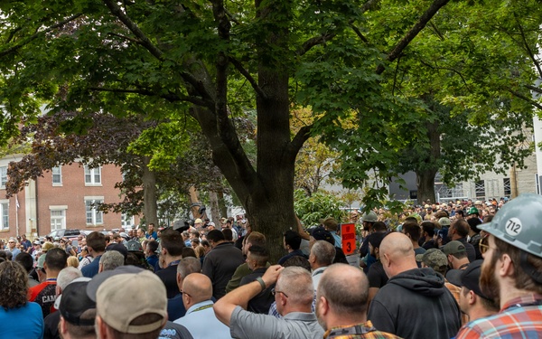 A large outdoor crowd of predominantly adult males has gathered in what appears to be a public square or plaza near brick institutional buildings. One individual in the foreground right is wearing a white hard hat marked with the number '712', suggesting union or trade worker affiliation. The crowd appears to be attending a rally, demonstration, or public gathering, with people facing toward a central focal point obscured by trees.